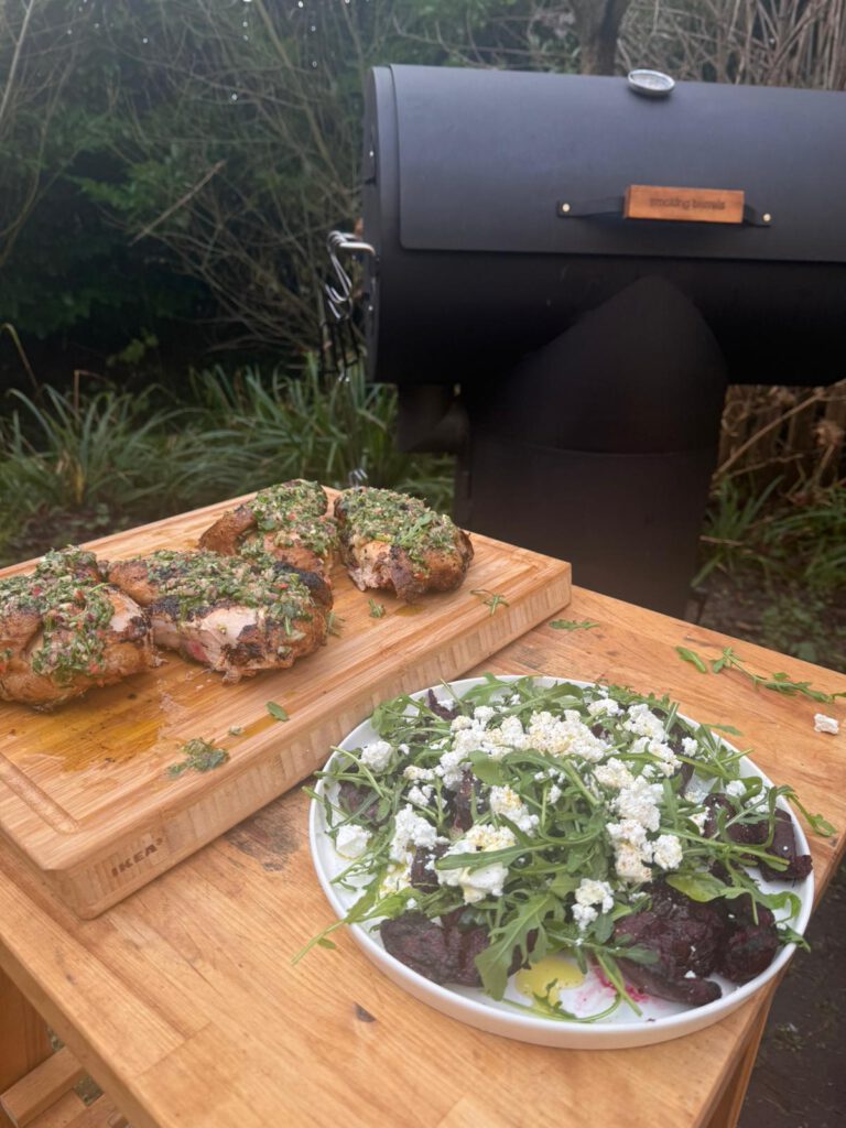 Gegrilde kip met chimichurri en een salade met rode biet, rucola en kaas, geserveerd op een houten tafel met The Barrel barbecue op de achtergrond.